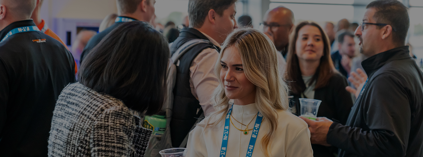 A group of people wearing conference lanyards converse and hold drinks in a busy indoor event space.