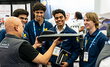 Several individuals stand around a table while a man showcases a drone in his hands.