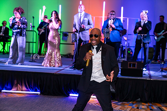 A band performs on stage with singers in formal attire and musicians playing instruments, illuminated by colorful lights. A man in a tuxedo sings with a gold microphone in the foreground.